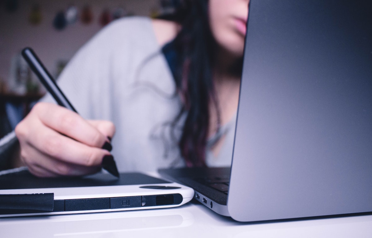 close-up of a person using a digital drawing tablet with a stylus while working on a laptop. The focus is on their hand and the tablet, with the person's face slightly blurred in the background.