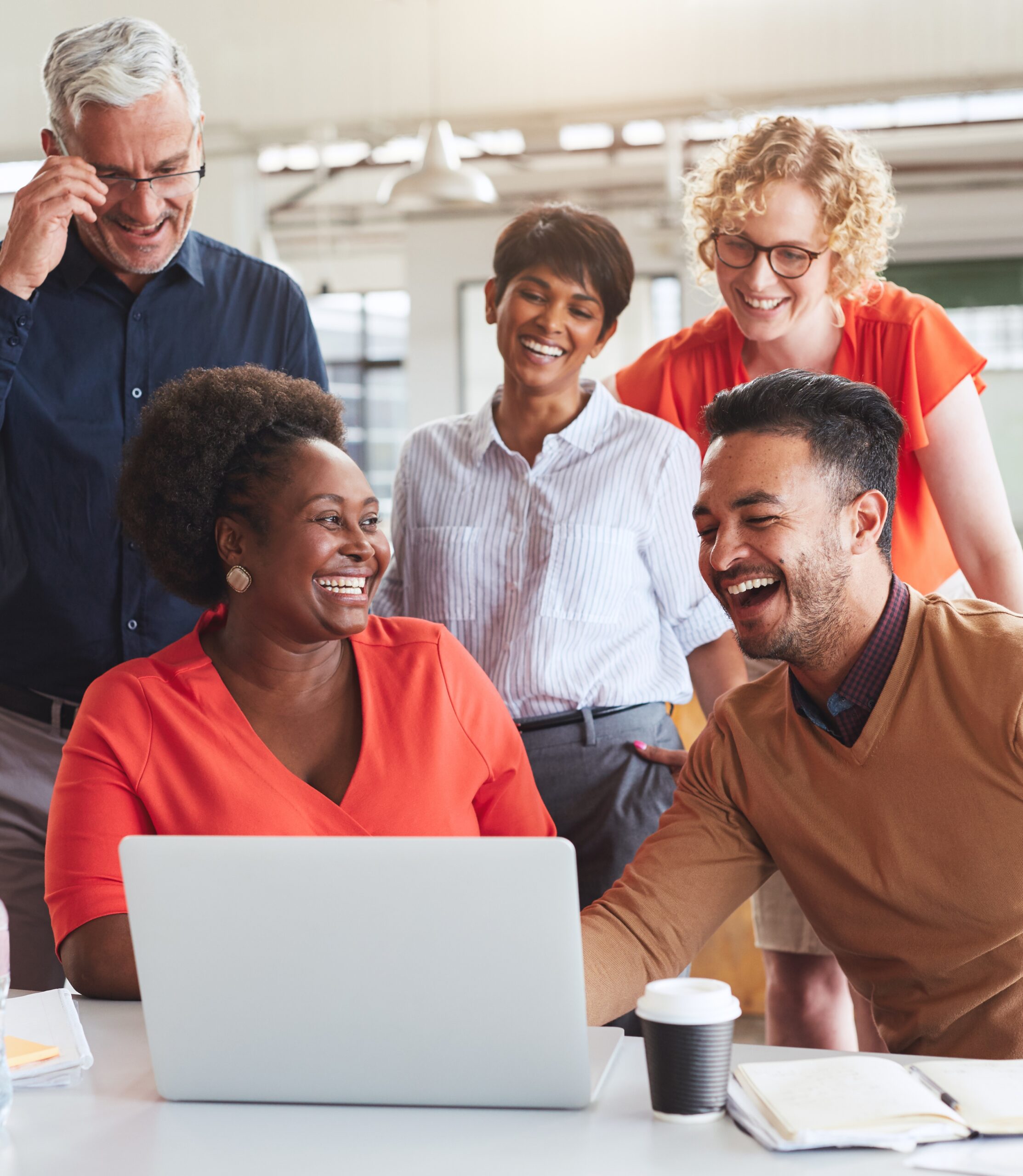 A group of smiling colleagues gathered around a laptop, engaging in a collaborative discussion in an office.