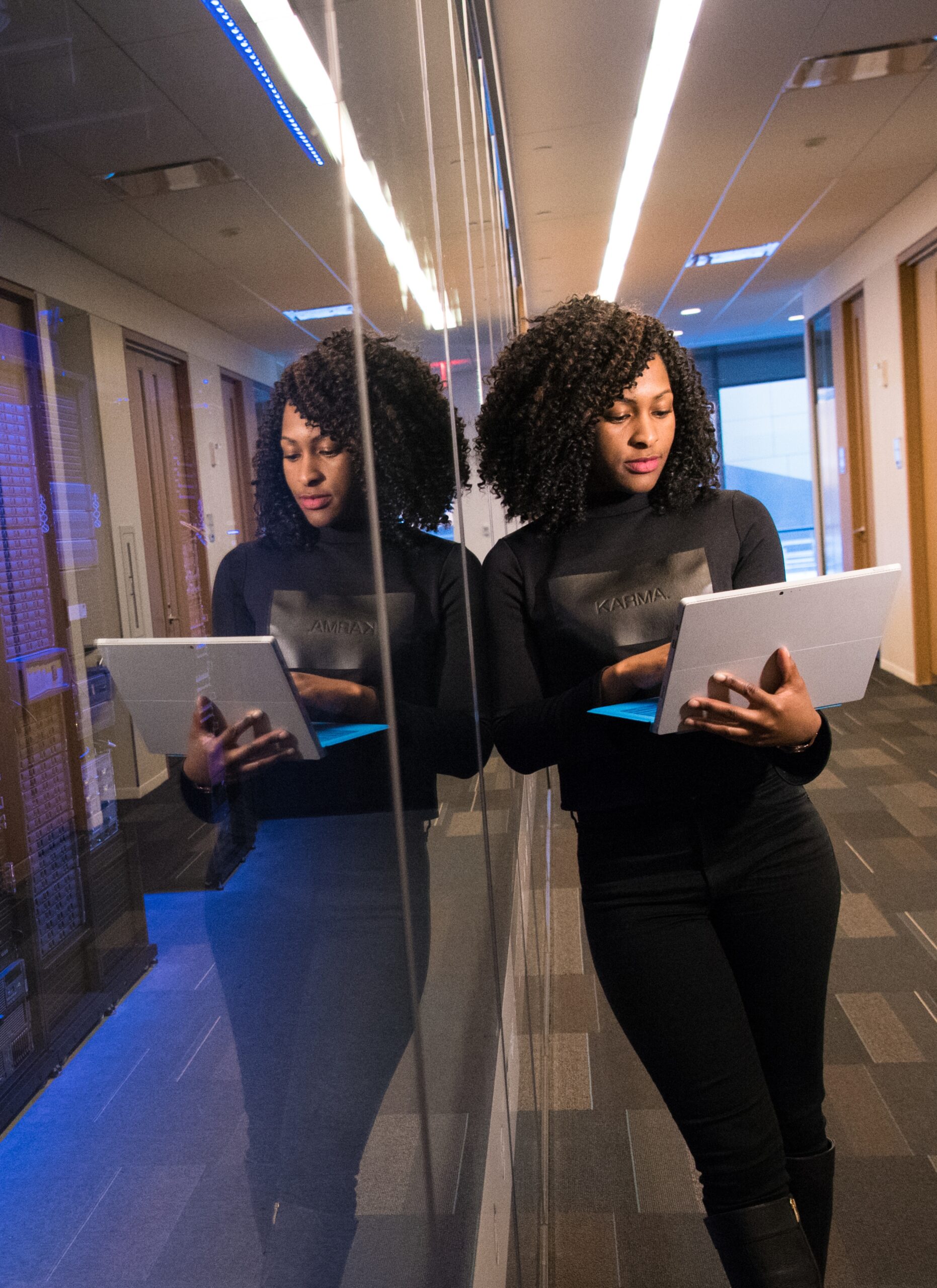 A woman in a reflective hallway, holding a tablet and working near server racks. Her reflection is visible on the glass panels of the racks.