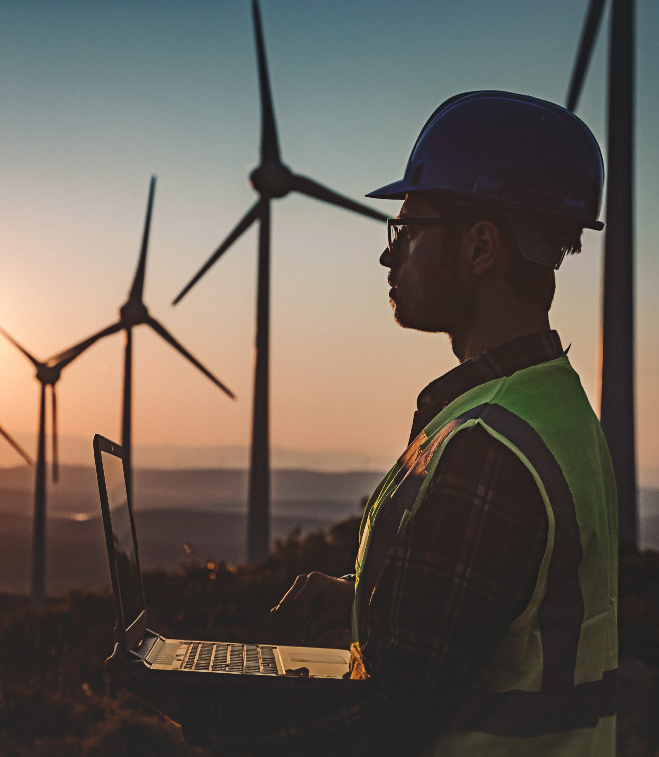 A worker in a hard hat and reflective vest using a laptop in front of wind turbines during sunset.