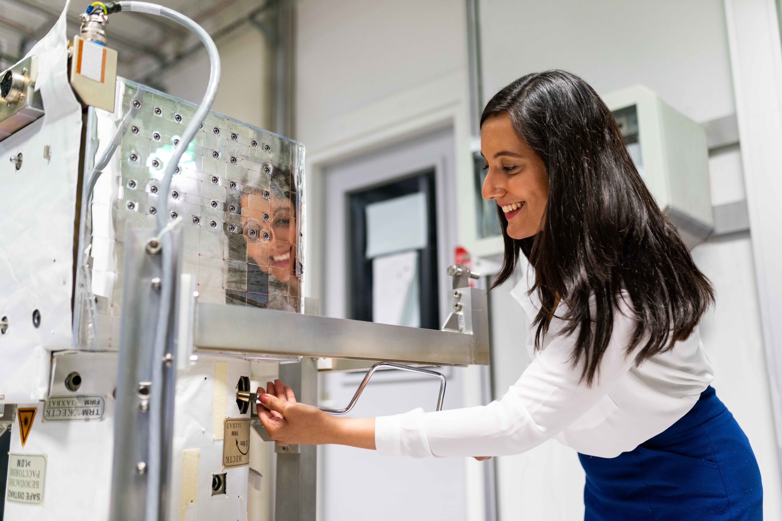 A woman smiling while working on a piece of laboratory equipment. Her reflection is visible on the metallic surface of the machine.