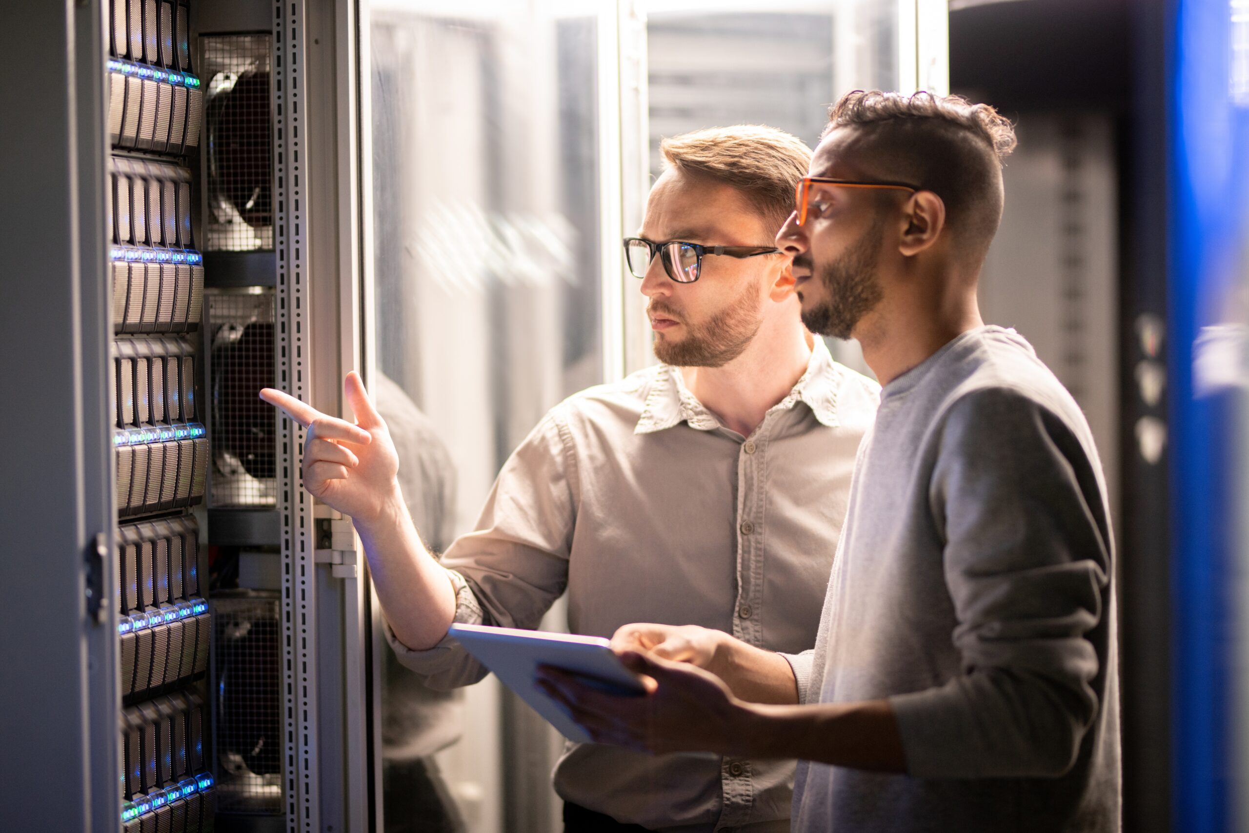 Two IT professionals in a server room discussing a server rack while one points at a component.