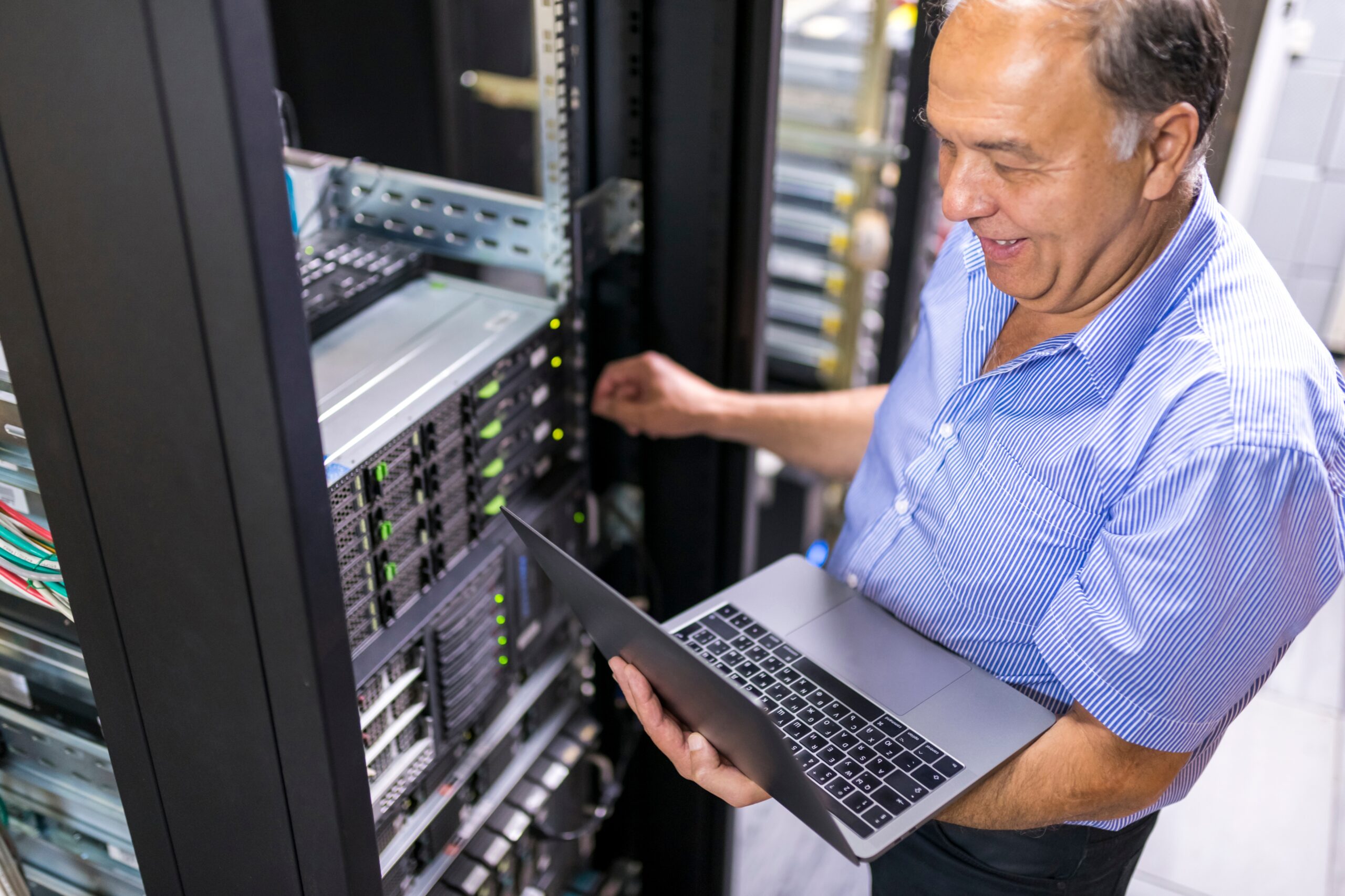 A senior IT professional inspecting server equipment while working on a laptop.