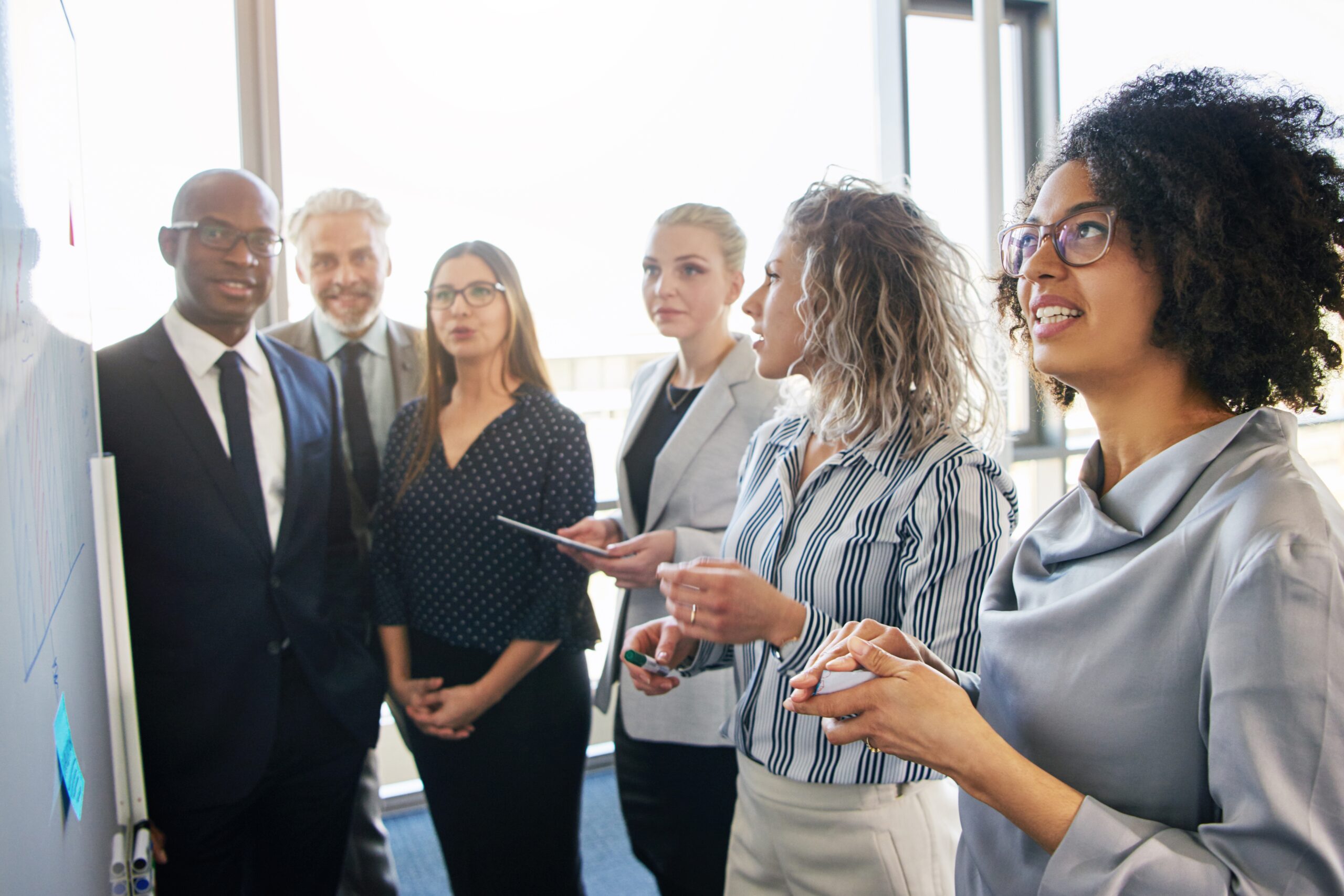 A diverse team of business professionals standing in front of a whiteboard, engaged in a discussion. The focus is on a woman speaking, with others actively listening.