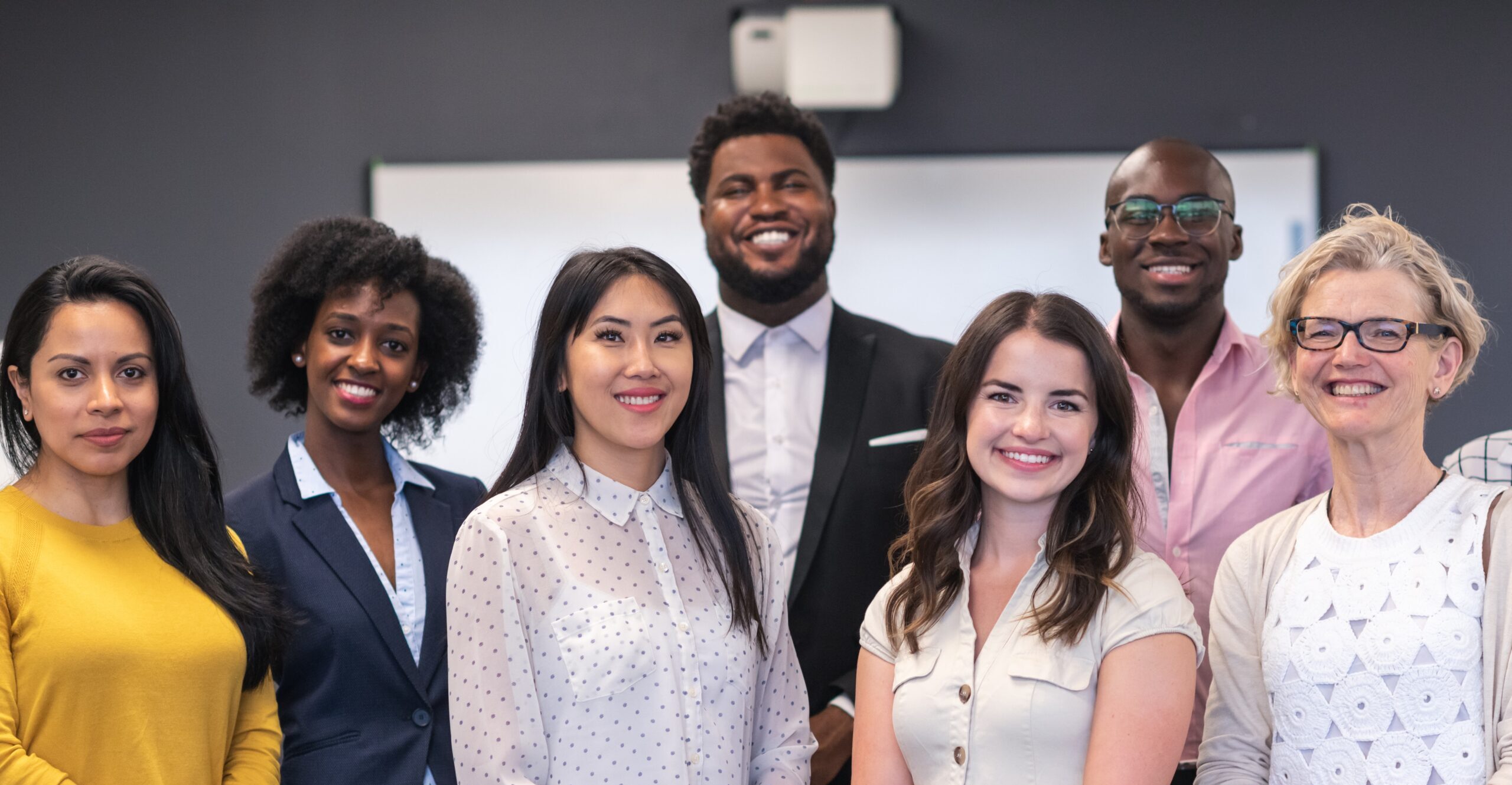 A diverse team of professionals, standing in a classroom setting, smiling and looking directly at the camera.