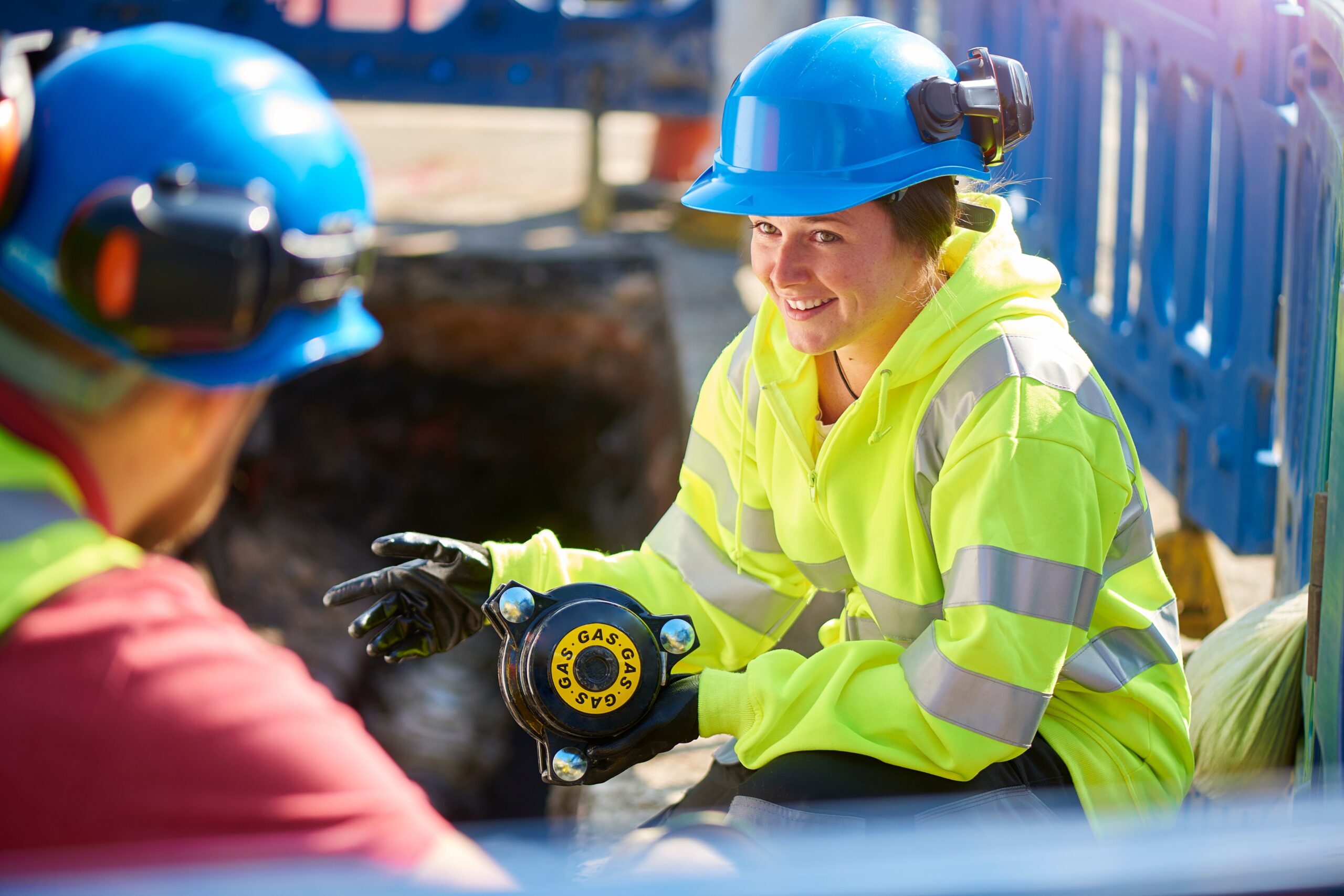 A female worker in a high-visibility jacket and hard hat kneeling on a construction site. She is holding a gas valve and smiling as she speaks with a colleague.