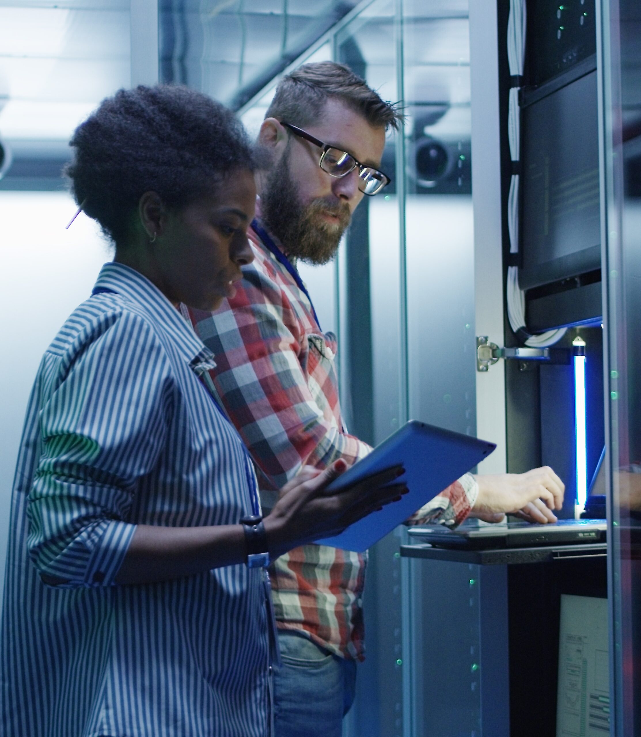 Two IT professionals working in a server room. A woman is holding a tablet, reviewing information, while a man operates a laptop. They are surrounded by server racks illuminated by blue lighting.