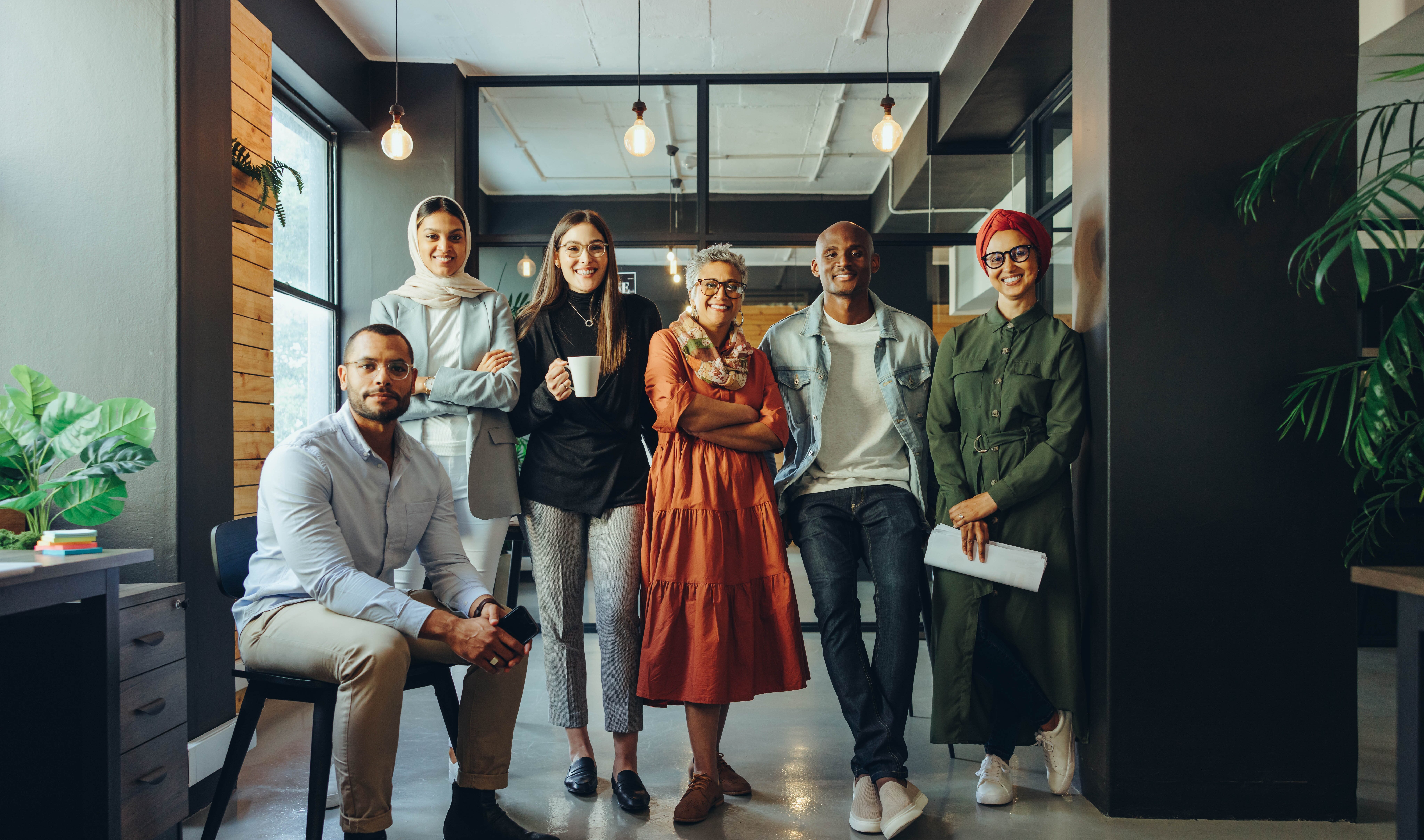 A diverse group of six professionals in a modern office, dressed in smart-casual attire, posing confidently with a warm, stylish background.