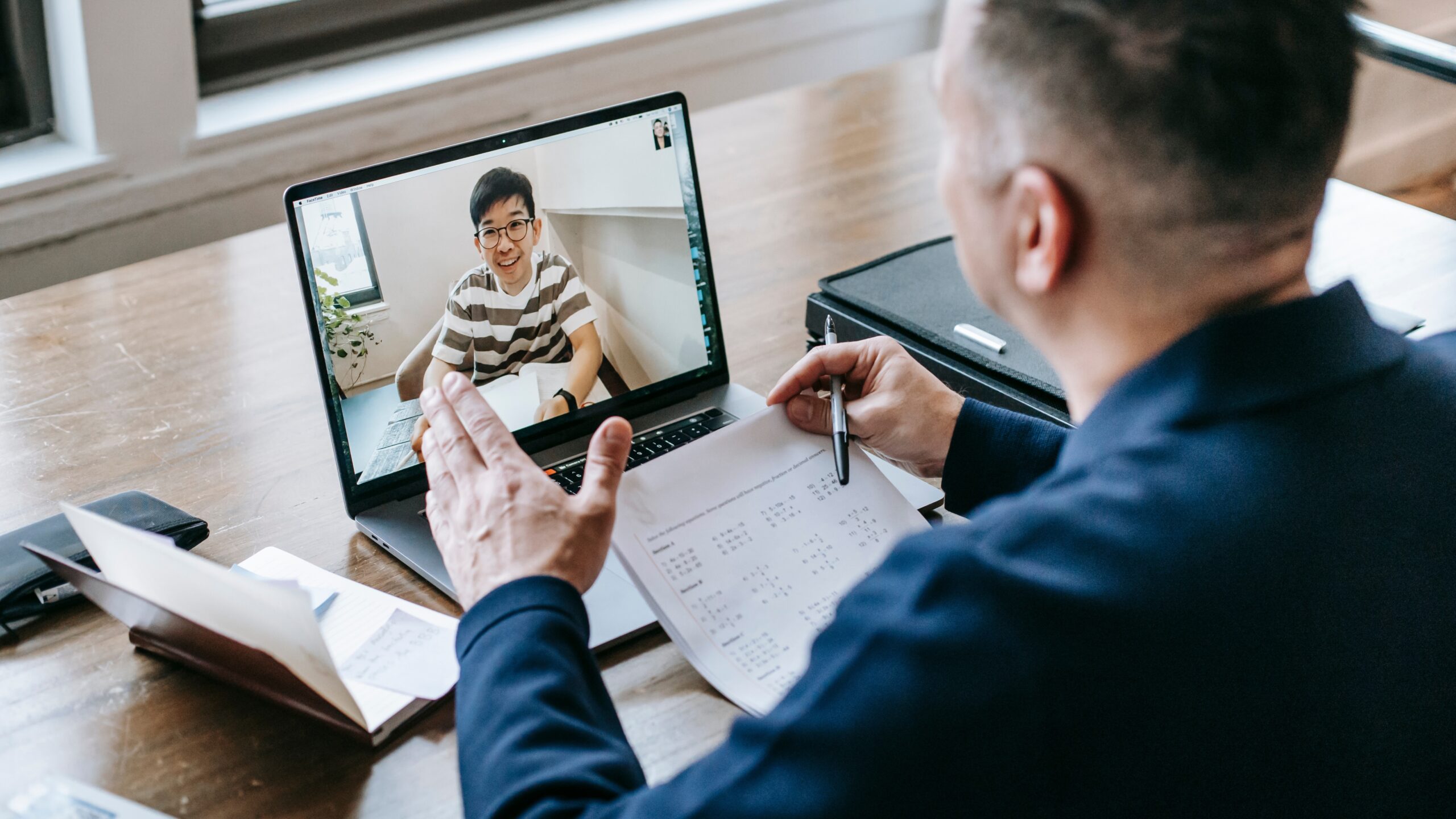 A man in a dark blazer gestures while speaking to a young man on a video call. The laptop screen shows the young man smiling, with a well-lit home setting in the background.