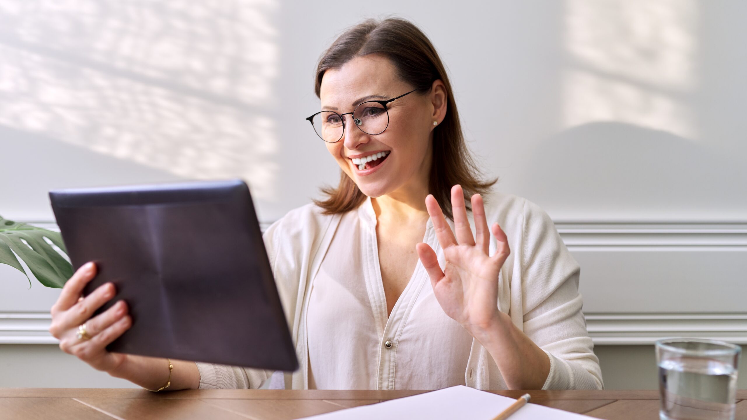 A woman wearing glasses and a light blouse waves at her tablet during a video call. Sunlight filters through the window behind her.