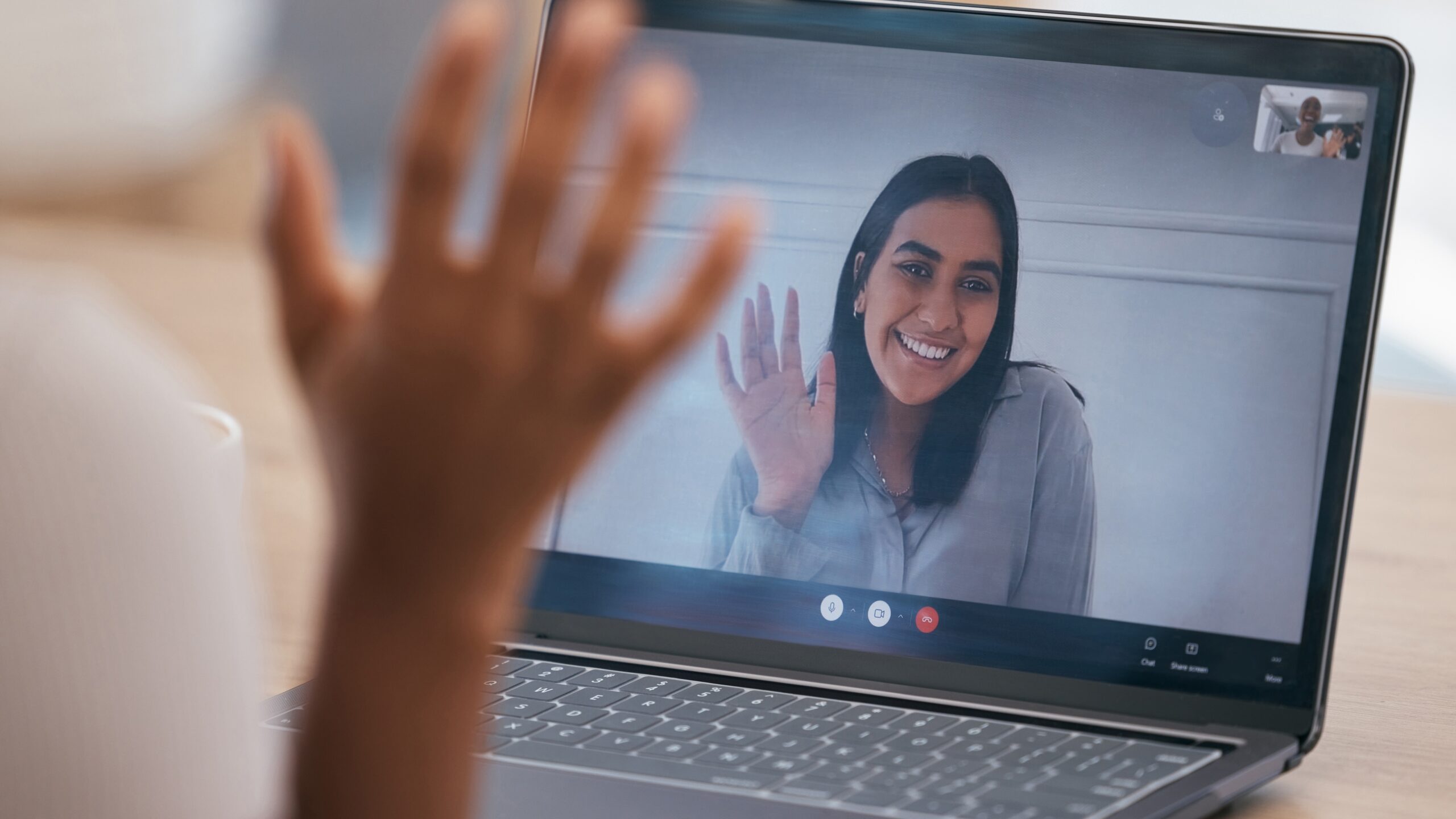 A close-up of a laptop screen showing a smiling woman waving during a video call. The person on the other end is out of focus, raising their hand.