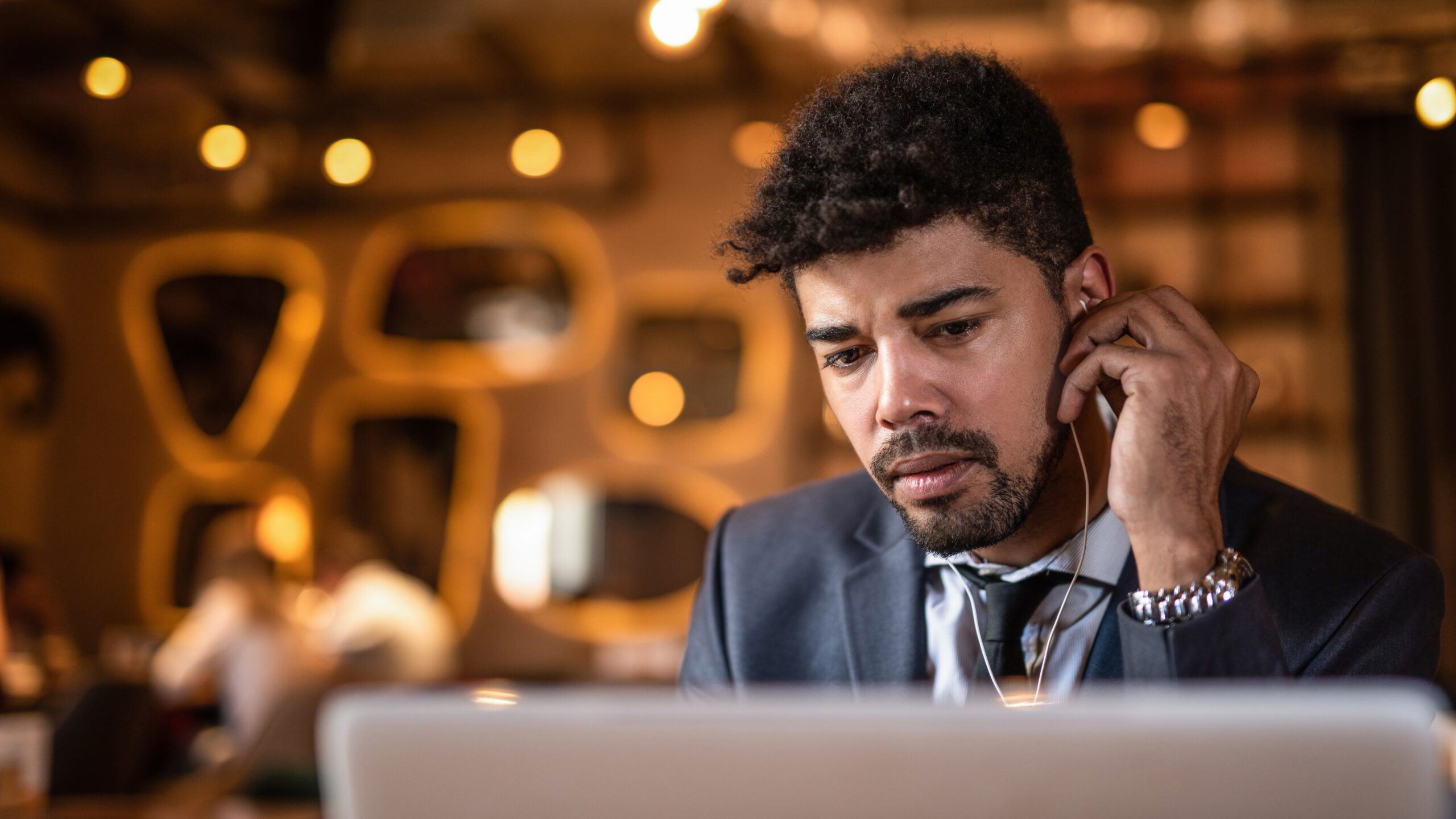A man in a suit with an earpiece looks focused while working on his laptop in a stylish, dimly lit café.