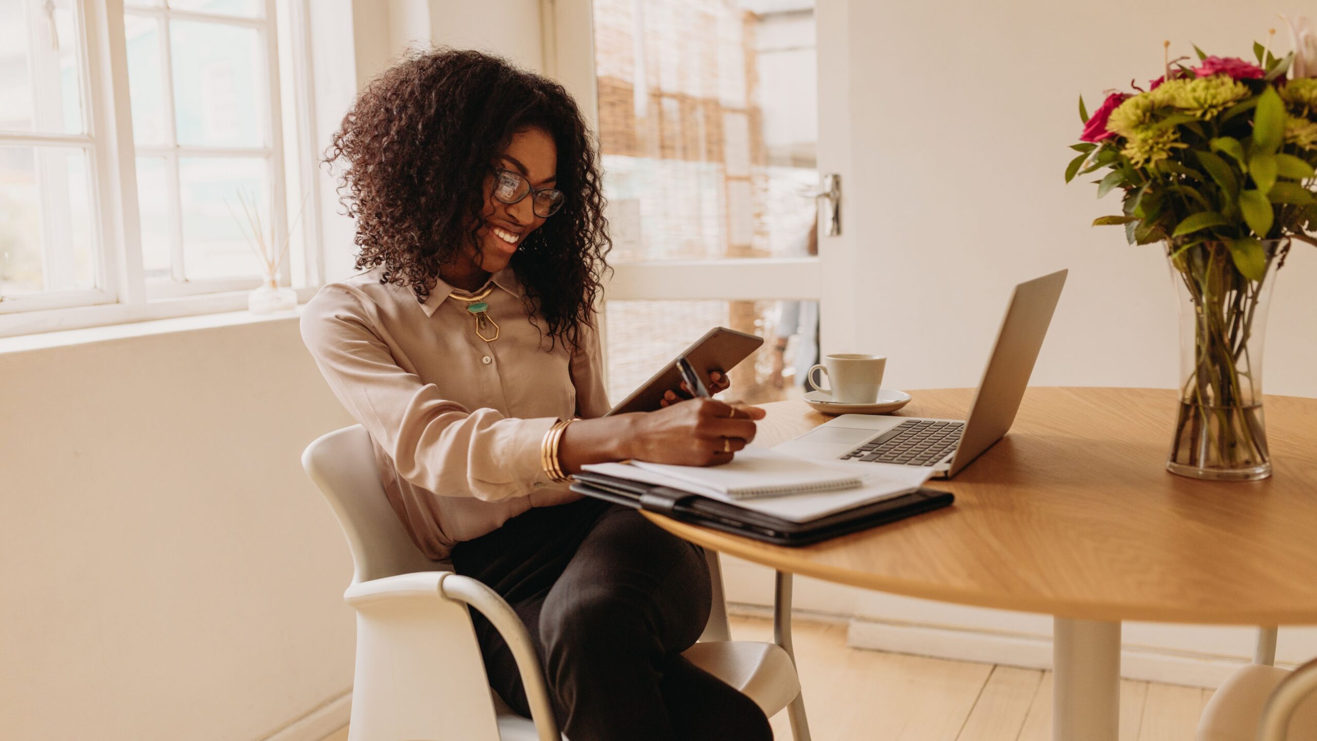 A woman sits at a table with a laptop, tablet, and notebook, smiling as she takes notes. A cup of coffee and a vase of flowers are beside her.