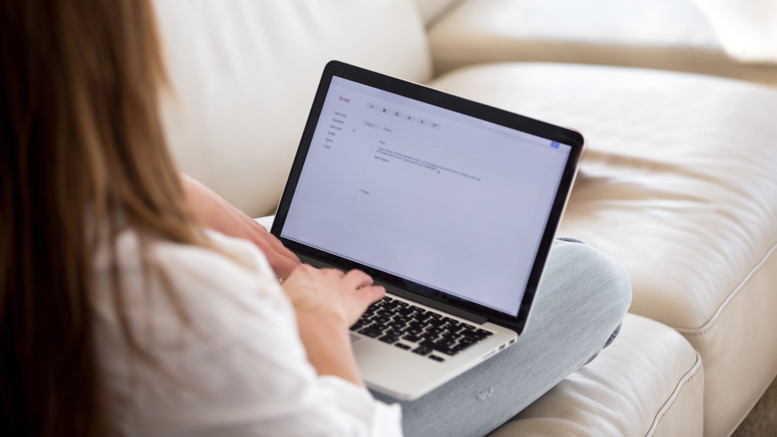 A woman sitting on a sofa types an email on her laptop, likely writing a cover letter or professional message. The screen displays a blank email composition window.