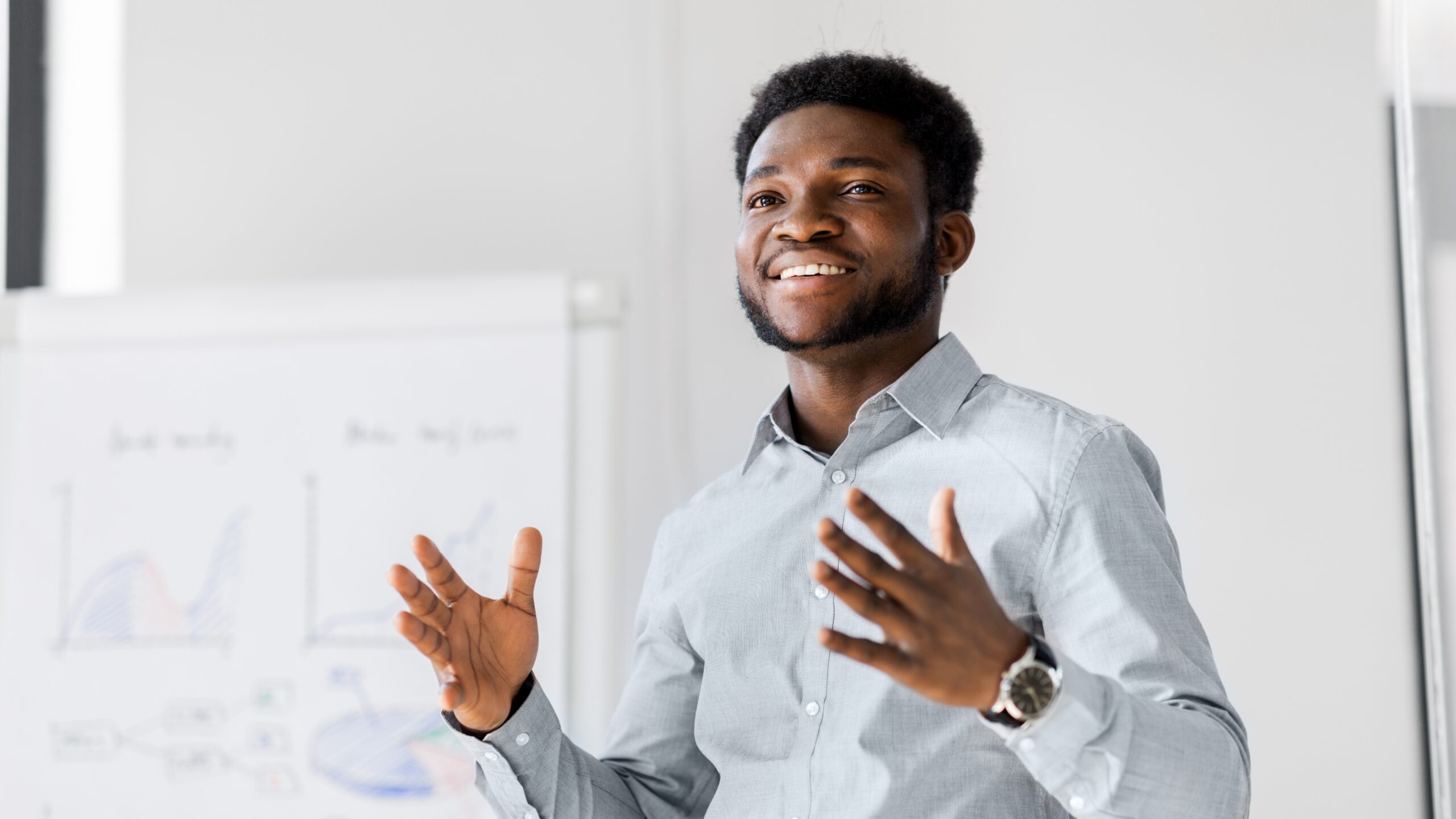 A confident man in a light grey shirt gives a presentation, gesturing with his hands while speaking. Behind him, a whiteboard displays charts and diagrams.