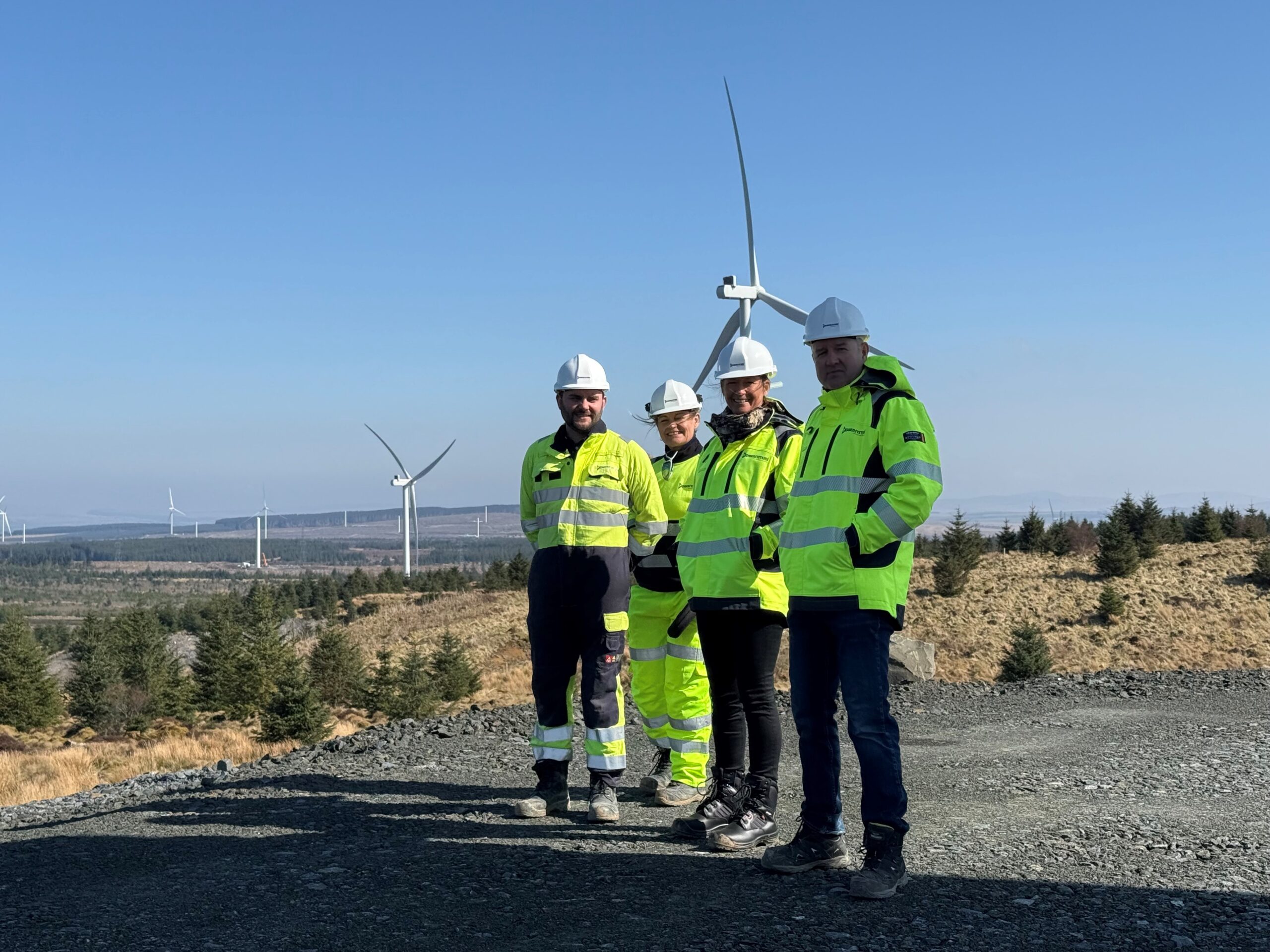 Four Powersystems workers in yellow high-visibility PPE and white hard hats stood in front of a field of wind turbines.