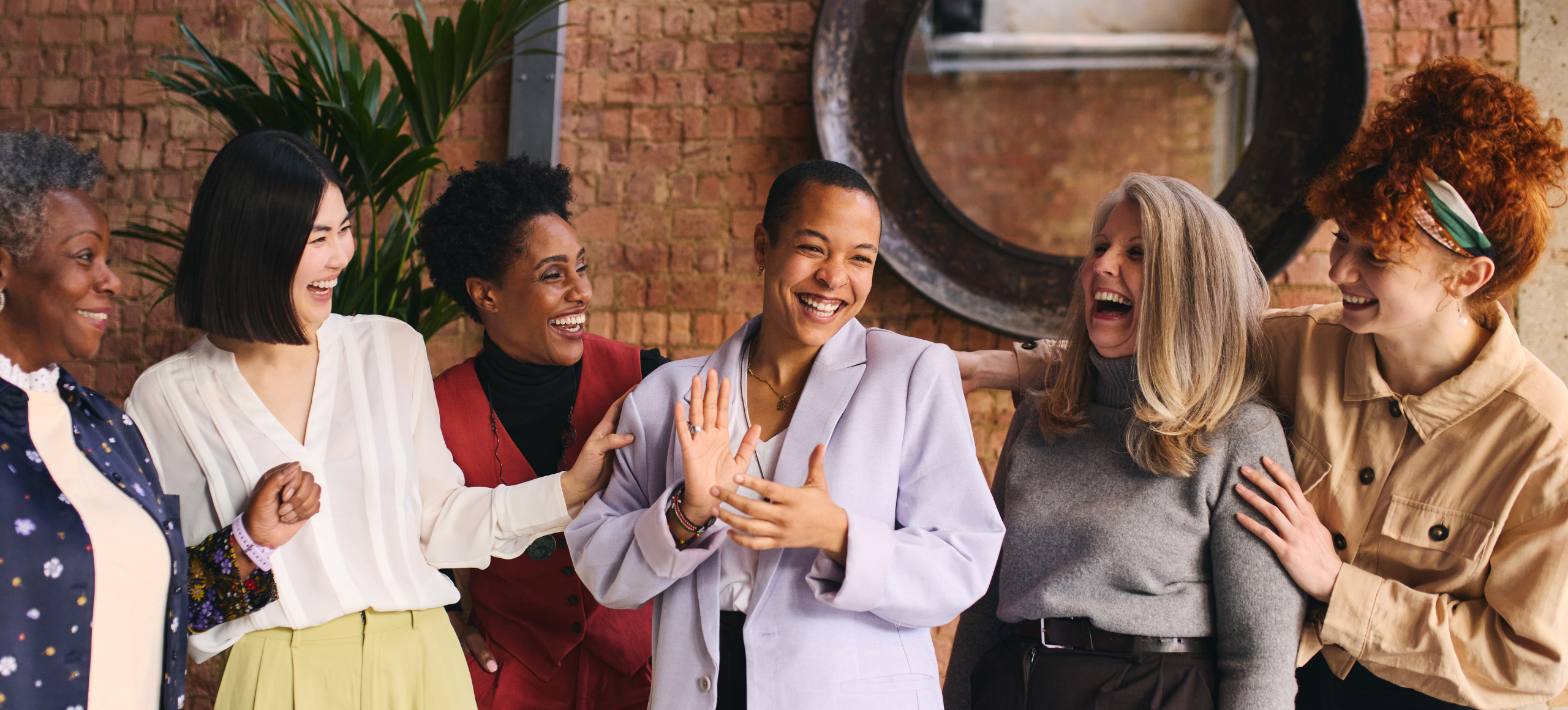 Six women of diverse ages and ethnicities stand together in a modern office with exposed brick walls and plants, smiling and laughing with their arms around each other.